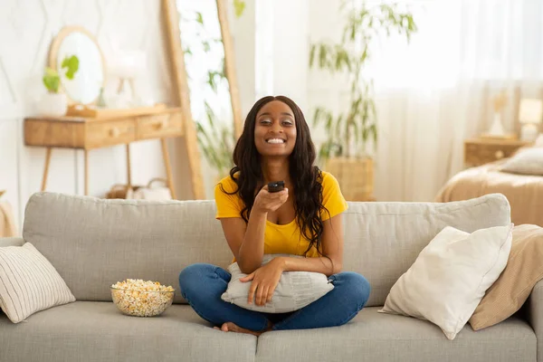 Μείνετε σπίτι χόμπι. Χαρούμενη μαύρη κυρία με τηλεχειριστήριο και ποπ κορν βλέποντας τηλεόραση στον καναπέ στο σαλόνι — Φωτογραφία Αρχείου