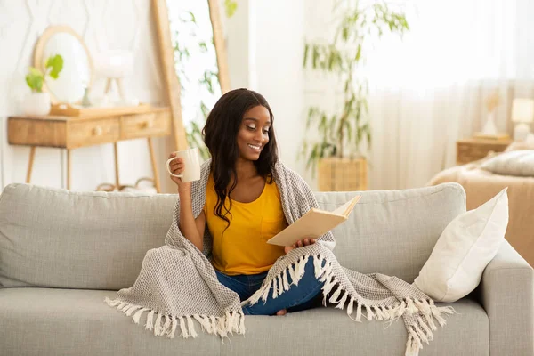 Güzel Afrikalı Amerikalı kadın elinde kahve fincanı ve kitap tutarken evdeki kanepede sıcak ekose altında oturuyor. — Stok fotoğraf