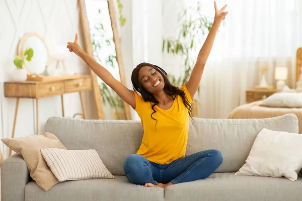 Heyecanlı kulaklıklı siyahi kadın müzik dinliyor ve ellerini evdeki kanepede kaldırıyor. — Stok fotoğraf