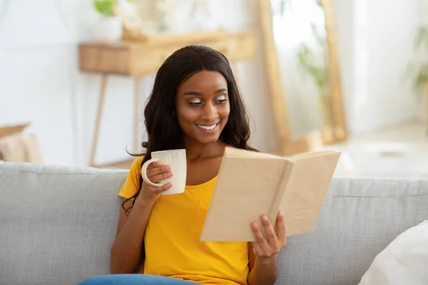 Μείνετε σπίτι δραστηριότητες αναψυχής. Υπέροχη νεαρή μαύρη γυναίκα ανάγνωση βιβλίο με φλιτζάνι ζεστό ρόφημα στο σαλόνι — Φωτογραφία Αρχείου