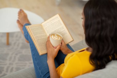 Afro-Amerikalı kadın, sabahları evde aromatik kapuçino ve büyüleyici bir kitapla tembellik ediyor.