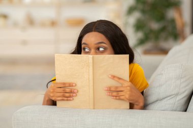 Ev eğlencesi konseptini bozma. Çekici Afrikalı Amerikalı kadın. Kapalı alandaki kanepede ilginç bir kitabın arkasına saklanıyor.