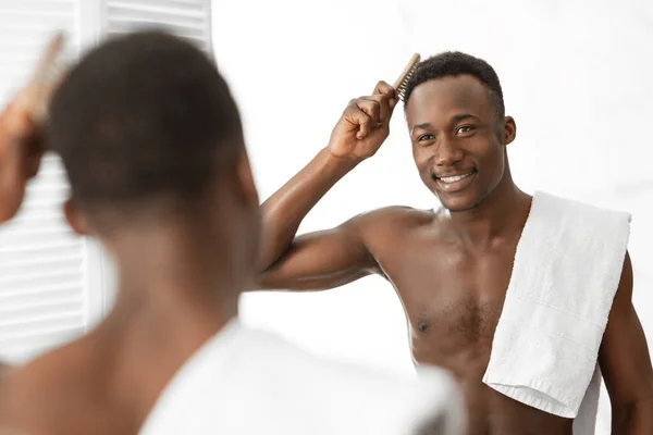 Sin camisa africano hombre cepillado pelo de pie cerca de espejo en cuarto de baño — Foto de Stock