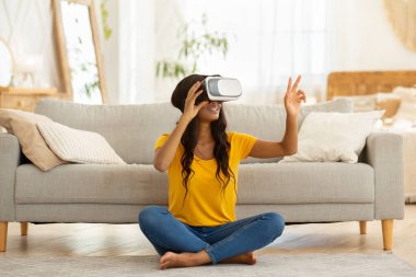 Evde sanal gerçeklik deneyimi sırasında VR kulaklıklı genç siyahi kadın havaya dokunuyor.