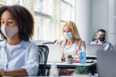 Ofisin içinde modern yönetici işyeri. Bin yıllık koruyucu maskeli sarışın kadın laptopta çalışıyor. Masadaki antiseptiğe odaklan.