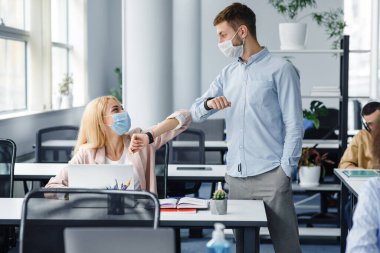Coronavirus salgını sırasında modern bir selamlama. Koruyucu maskeli genç adam ve kadın ofisin içinde birbirlerine dirsekleriyle dokunuyorlar.