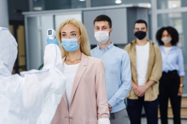 İş yerinde sıcaklık kontrol testi ve sosyal uzaklık. Koruyucu giysili adam sabahları temas olmayan termometreyle çok uluslu çalışanları kontrol ediyor.