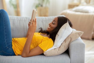 Haftasonu evde kalıp hobi edin. Açık kitabı olan genç siyahi kadın rahat koltukta uzanır.