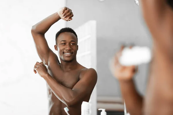 Feliz hombre negro aplicando desodorante de pie cerca del espejo en el baño — Foto de Stock