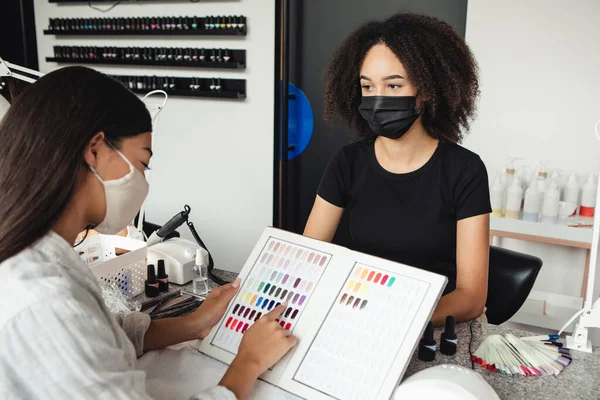 Werken in nagelsalon tijdens COVID-19 epidemie. Aziatisch klant dame in beschermende masker kiezen kleur voor nagels, Afrikaans amerikaanse vrouw kijkt naar klanten — Stockfoto