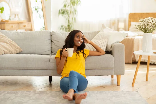 Pasando un fin de semana tranquilo en casa. Milenial dama negra disfrutando de su café en el suelo cerca del sofá en el interior —  Fotos de Stock