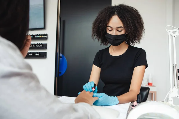 Work with client after quarantine in salon. African american woman in protective mask covering nails of asian client with polish in studio during covid-19 epidemic