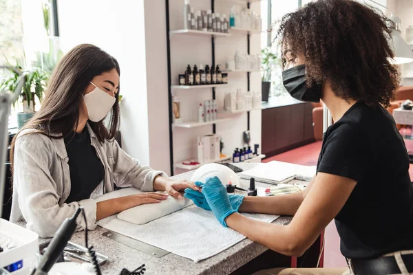 Terug aan het werk na quarantaine en roddels in de nagelsalon. Afrikaans amerikaanse vrouw in beschermende masker bestanden nagels met elektrische apparaat naar cliënt — Stockfoto