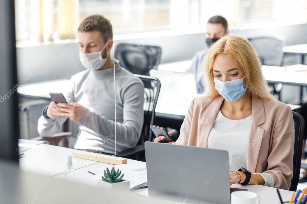 Modern gadgets for working with clients in office. Millenial man in mask typing on smartphone, lady works in laptop at workplace with protective glass