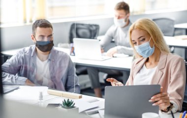 Coronavirüs salgını sırasında projeyle ilgili görüşmeler ve tartışmalar. Koruyucu maskeli genç adam ve kadın laptopa bakar ve iş yerindeki cam levhalarla iletişim kurarlar.