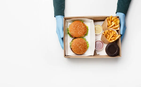 Lunch dla dwojga podczas epidemii koronawirusów. Kelner w rękawiczkach trzyma pudełko z zestawem hamburgerów i cheeseburgerów, frytki w opakowaniach ekologicznych, ketchup i sos w pojemnikach i kawie — Zdjęcie stockowe