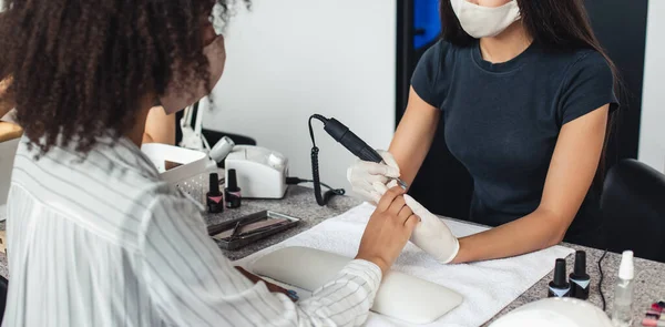 Existência de pequenas empresas no encerramento da COVID-19. Mestre de manicure profissional em máscara protetora faz procedimento de manicure com dispositivo elétrico para cliente feminino afro-americano à mesa — Fotografia de Stock