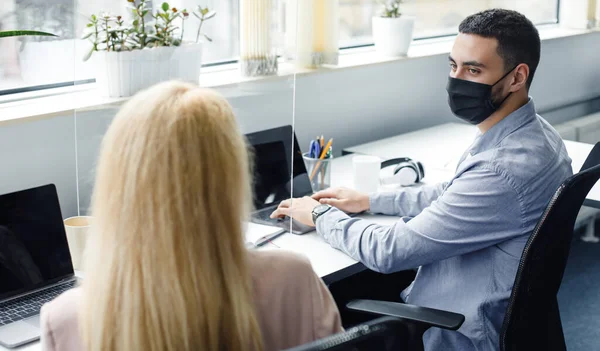 Local de trabalho moderno no escritório de coworking após a quarentena vívida. Cara e senhora em máscaras sentado à mesa e trabalhando em laptop, falando, através de vidro protetor — Fotografia de Stock