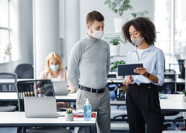 Boss and manager discussion of work and social distance. African american woman in protective mask shows tablet to male worker near table with laptop