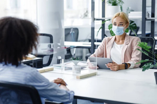 Praten met de baas of interviewen tijdens de coronavirusepidemie. Jonge vrouw in beschermende masker met tablet communiceert met Afrikaanse Amerikaanse dame door middel van beschermende glas — Stockfoto