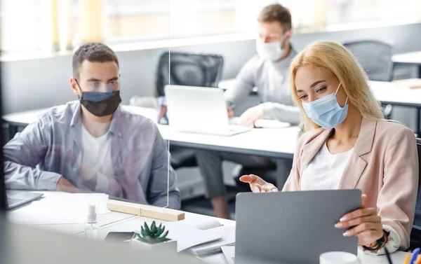 Bijeenkomst en bespreking van het project tijdens de coronavirusepidemie. Jonge man en vrouw in beschermende maskers kijken naar laptop en communiceren via glazen bord op de werkplek — Stockfoto