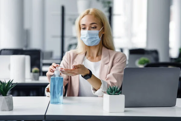 Безопасность и охрана здоровья на работе в офисе. Тысячелетняя женщина в защитной маске с умными часами использует антисептик на рабочем месте с ноутбуком для дезинфекции рук — стоковое фото