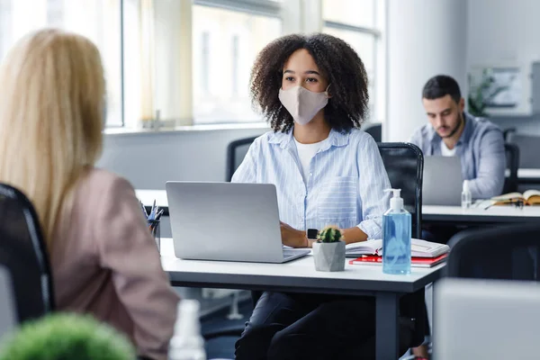 Work with client after quarantine and social distancing. Friendly african american woman in protective mask talking to european lady, sitting at workplace with laptop — Stock Photo, Image