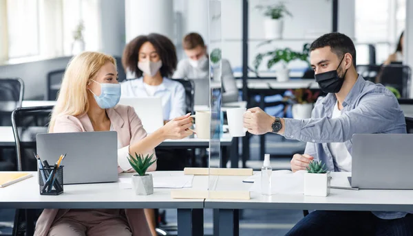 Modern ofiste kahve molası ve işçilerin sağlığını korumak. Koruyucu maskeli genç adam ve kadın ofis içi cam panolardan bardakları tokuşturuyorlar.