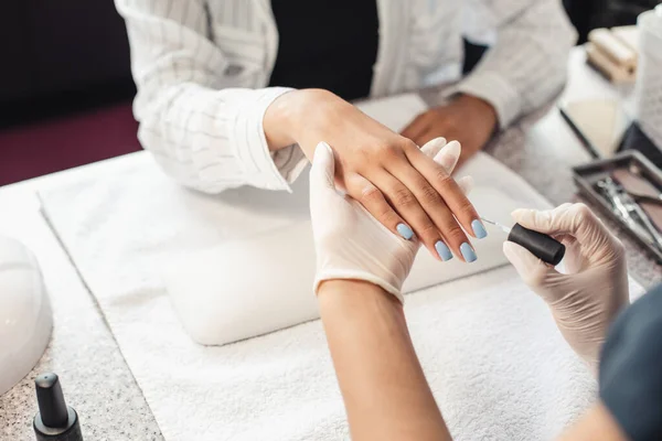 Reabertura do salão de beleza e cuidados com as unhas após a quarentena. Mestre de manicure em luvas de borracha pinta unhas com gel azul polonês para Africano americano senhora cliente — Fotografia de Stock