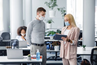 Yeni normal, sosyal mesafeli ve çalışan iletişimi olan bir patron. Koruyucu maskeli iş kadını erkeklere tablet gösteriyor. İş yerinin yakınında laptoplu ve antiseptik.