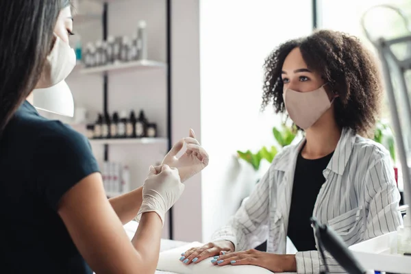 Profissional mestre manicure e distanciamento social. Mulher asiática em máscara protetora colocar luvas de borracha para começar a trabalhar com unhas de senhora americana africana — Fotografia de Stock