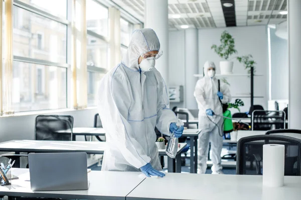 Disinfection of premises after work during covid-19 epidemic. People in hazmat suits, goggles and masks clean furniture with chemicals in office interior — Stock Photo, Image