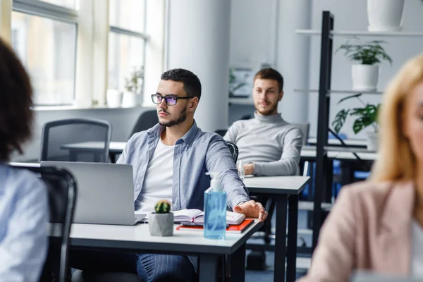 Crear ideas para trabajar después de la cuarentena. Joven hombre atractivo en gafas mira por la ventana, sentado en la mesa con el ordenador portátil, planta en maceta y antiséptico — Foto de Stock