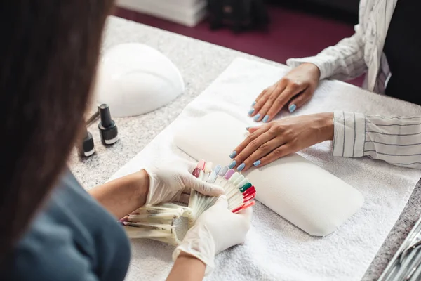 Schoonheidsbehandeling in moderne salon na quarantaine. Professionele meester in rubber handschoenen met behulp van vernissen monsters voor klant, Afrikaans-Amerikaanse vrouw zet haar handen op tafel — Stockfoto