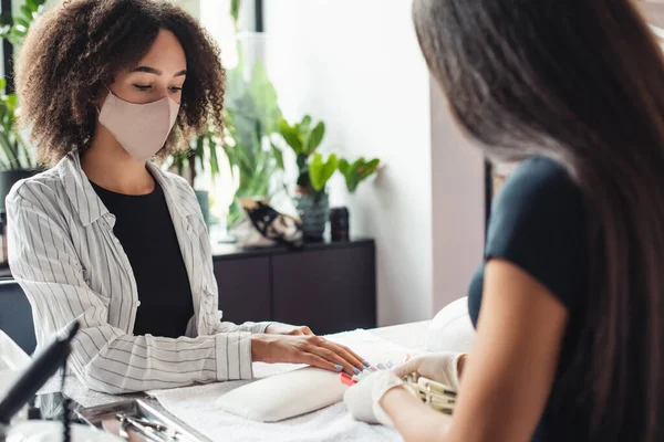 Manicure and pedicure salon, covid-19 and social distance. Master in rubber gloves and african american young woman client in protective mask choose color for manicure