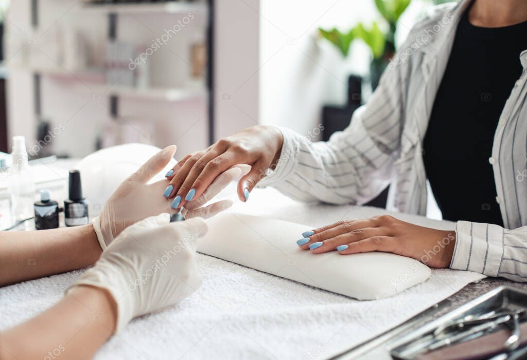 We are open, reopening of beauty salon. Female master in rubber gloves cover blue polish to african american lady client nails in studio interior during coronavirus epidemic