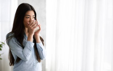 Rahatlamış Asyalı kadın çay içiyor ve evdeki pencereden manzaranın tadını çıkarıyor.