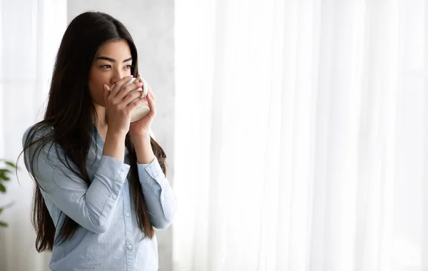Relajada mujer asiática bebiendo té y disfrutando de la vista desde la ventana en casa —  Fotos de Stock