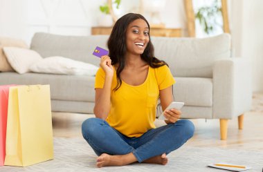 Siber alışveriş konsepti. Akıllı telefonu ve kredi kartı olan Afrikalı Amerikalı bir kadın evde internetten bir şeyler sipariş ediyor.