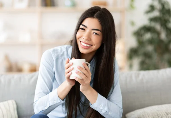 Mujer coreana sonriente bebiendo café mientras descansa en el sofá en casa — Foto de Stock