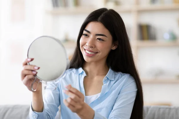 Mulher asiática bonita segurando espelho de ampliação em casa, olhando para o seu reflexo — Fotografia de Stock