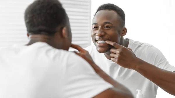 Africano hombre chequeando dientes sonriendo a espejo de pie en cuarto de baño — Foto de Stock