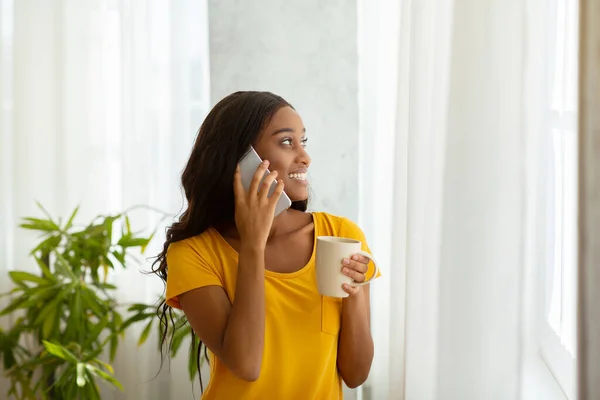 Великолепная афроамериканка, стоящая у окна с чашкой кофе и общающаяся по смартфону дома — стоковое фото