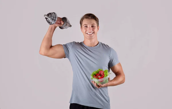 Hälsosam kost och motion. Glad vit kille lyfta hantel och hålla grönsakssallad på ljus bakgrund — Stockfoto