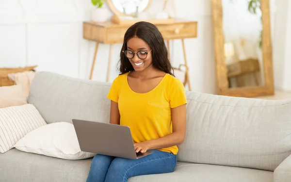Ritratto di giovane donna nera che utilizza il computer portatile per lavoro o istruzione sul divano a casa — Foto Stock