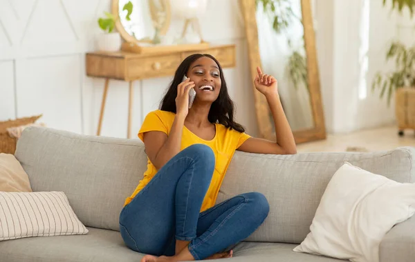 Mulher afro-americana encantadora falando no smartphone e rindo no sofá em casa — Fotografia de Stock