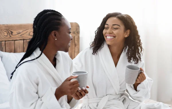 Gelukkige vriendinnen in badjassen zittend op bed en koffie drinkend — Stockfoto