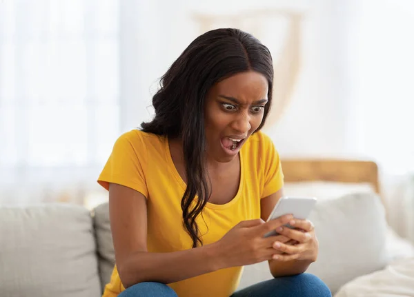 Jovem senhora negra recebendo notícias terríveis, com raiva de seu telefone celular em casa — Fotografia de Stock