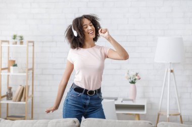 Hayali mikrofonda şarkı ve müzikal mola. Covid-19 salgını sırasında oturma odasının iç kısmında kulaklıklı neşeli Afrikalı Amerikalı kadın şarkı söylüyor.