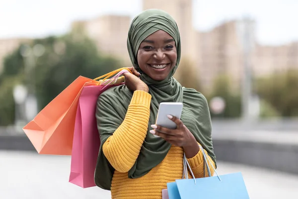 Çevrimiçi bankacılık. Akıllı Telefonlu Neşeli Zenci Müslüman Kadın ve Açık havada Alışveriş Çantaları — Stok fotoğraf